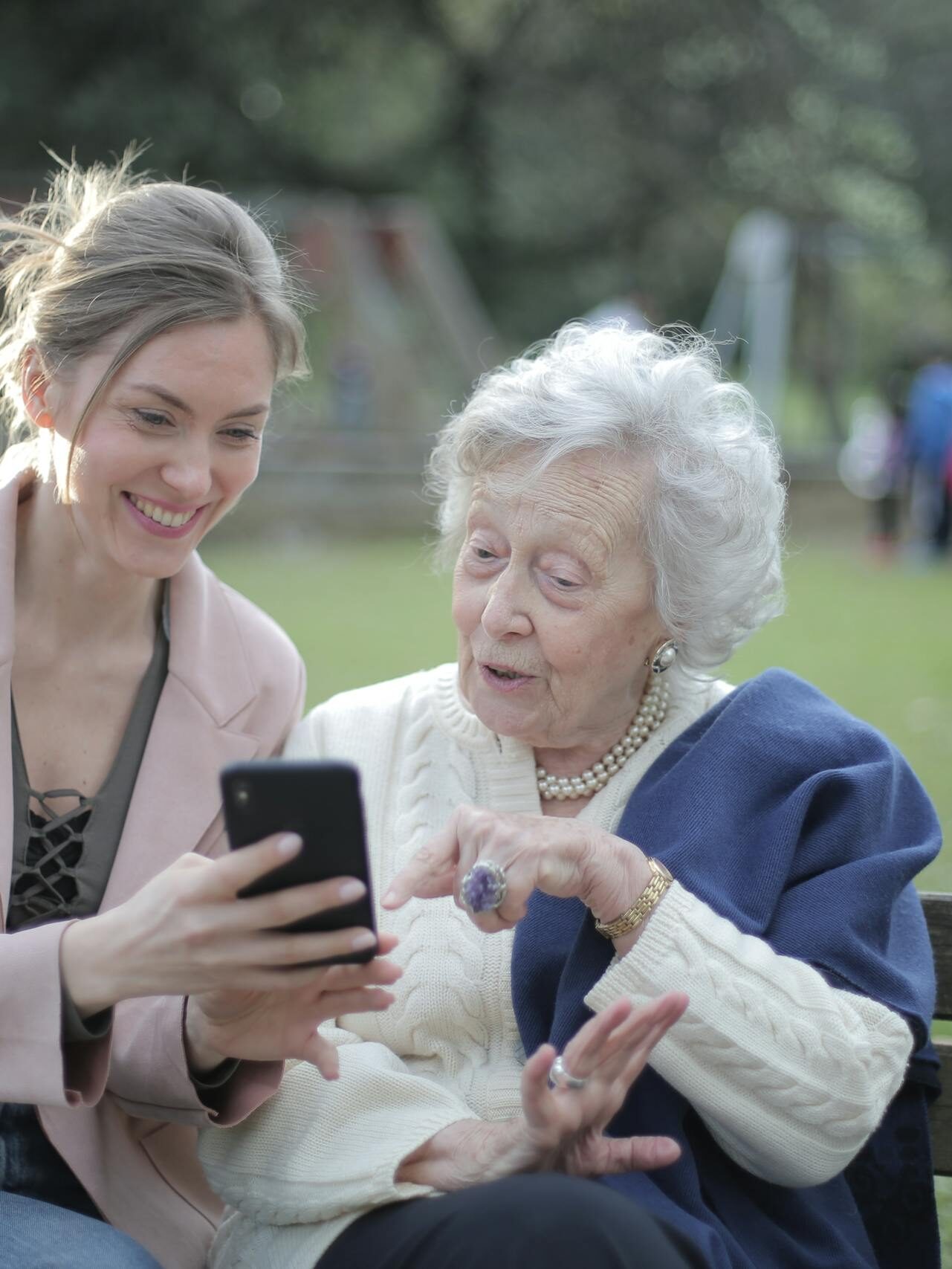 Delighted female relatives sitting together on wooden bench in park and browsing mobile phone while learning using